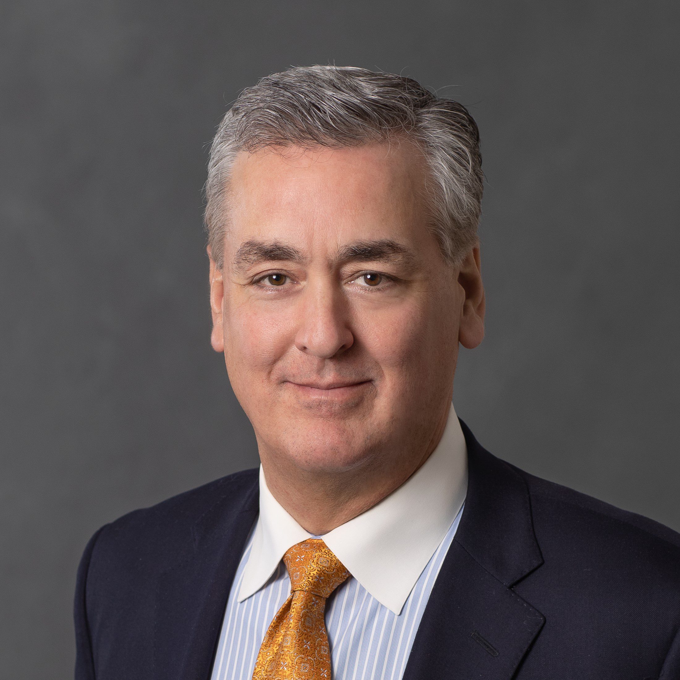 John Valente III of the Valente Law Group in a dark suit, pin striped shirt, and patterned orange tie, smiling against a gray background.