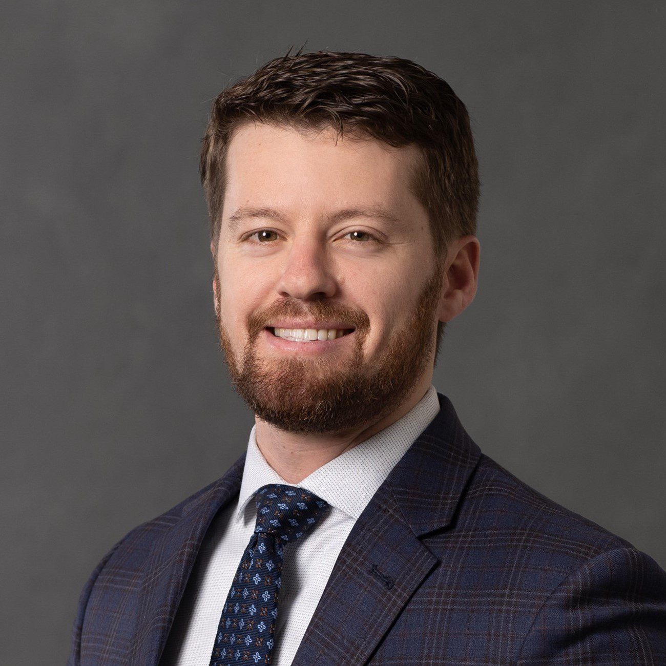 Joseph Chandlee of the Valente Law Group in a dark plaid suit, white shirt, and patterned blue tie, smiling against a gray background.