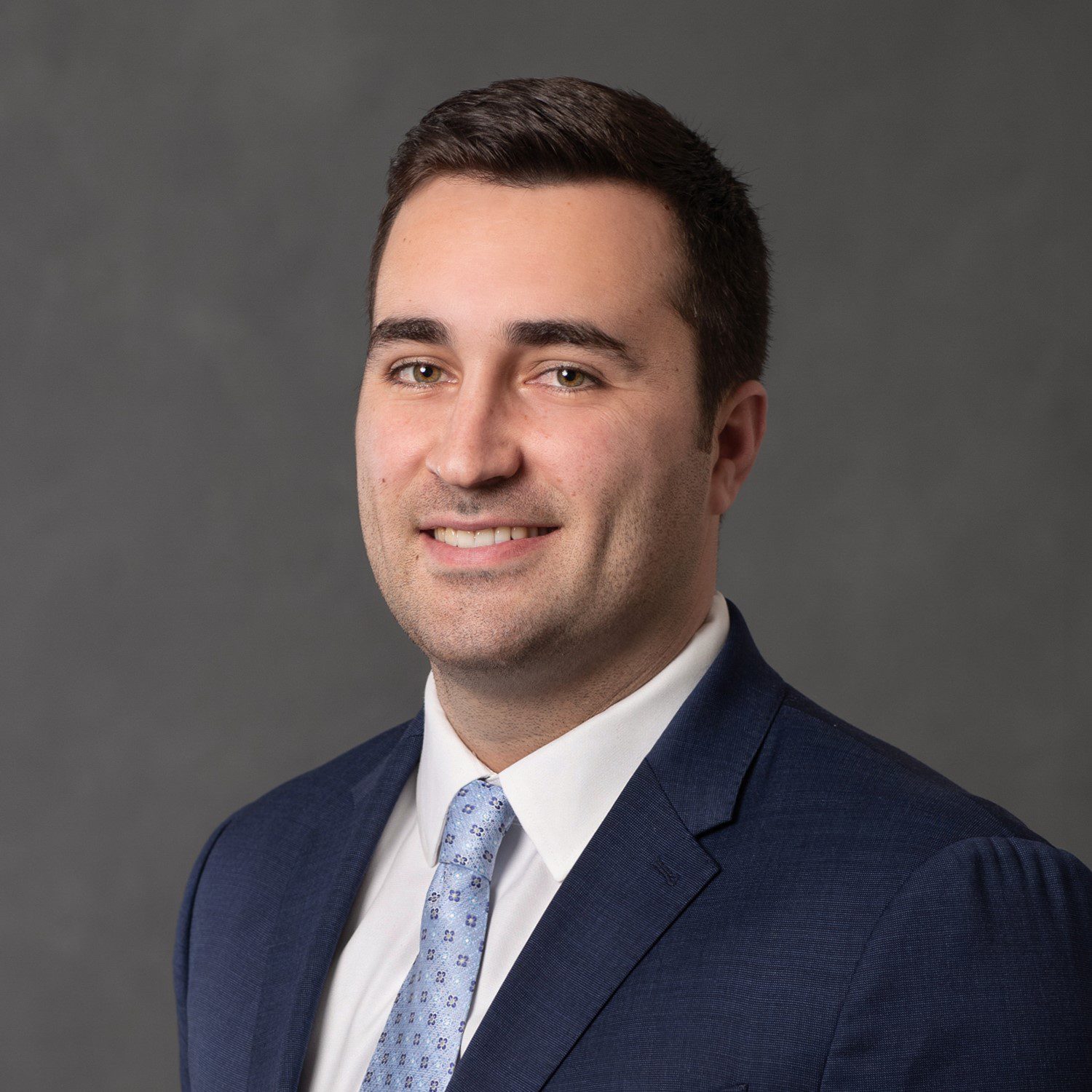 Michael Mancuso Jr. of the Valente Law Group in a dark blue suit, white shirt, and light blue patterned tie, smiling against a gray background.
