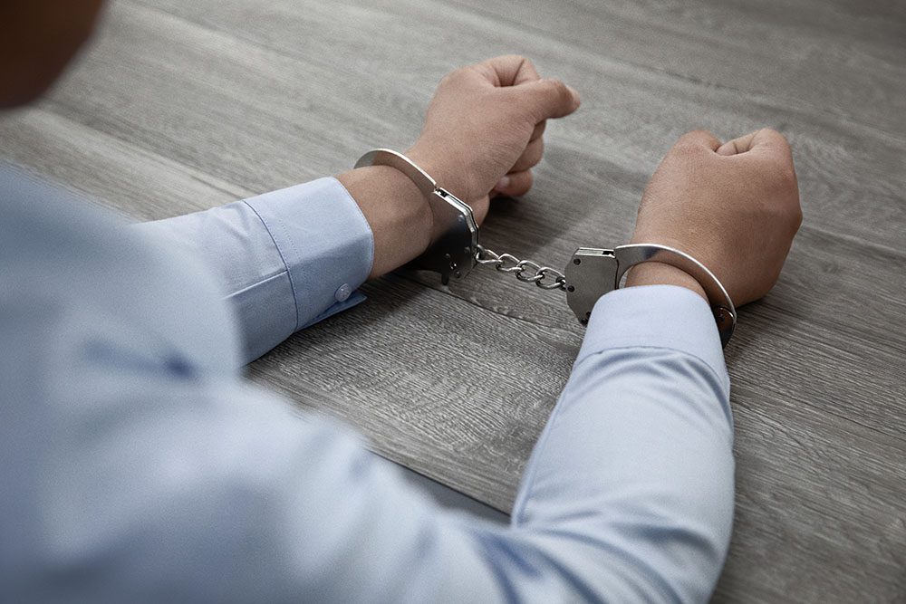 A person's hands in handcuffs, resting on a wooden surface. The individual is wearing a light blue dress shirt.